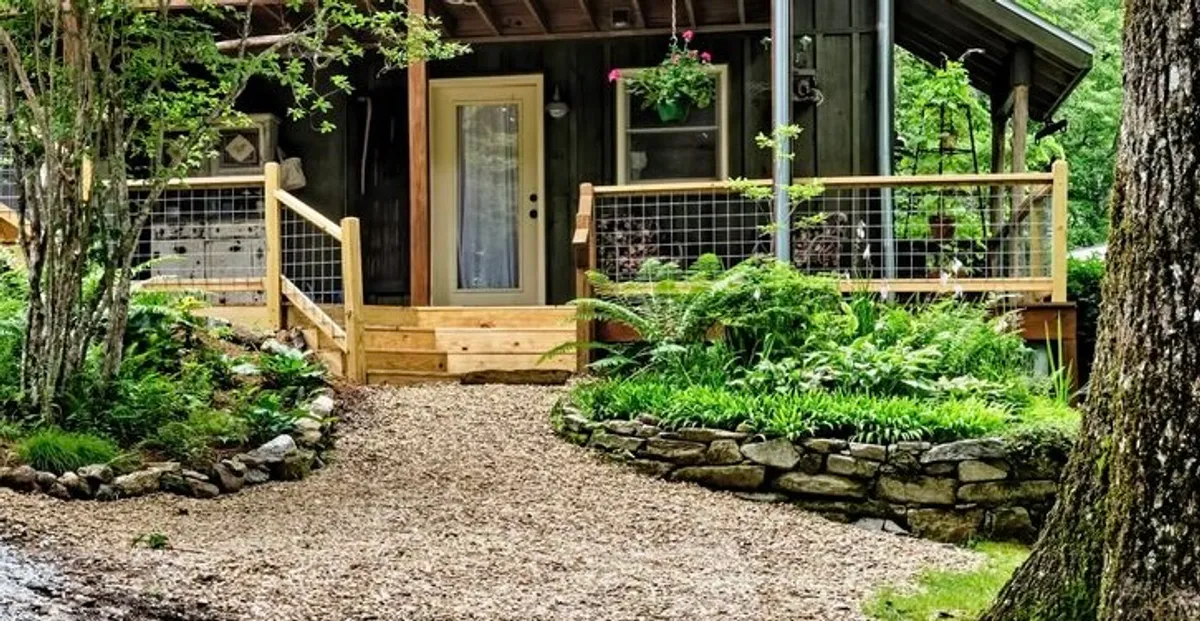 The image displays the inviting exterior of 'The Cozy Cub' apartment, featuring a welcoming front door at the top of rustic wooden steps. Lush ferns and seasonal blooms line a gravel pathway and stone border, all softly illuminated by morning sunlight filtering through the surrounding trees, creating a serene and peaceful entrance.