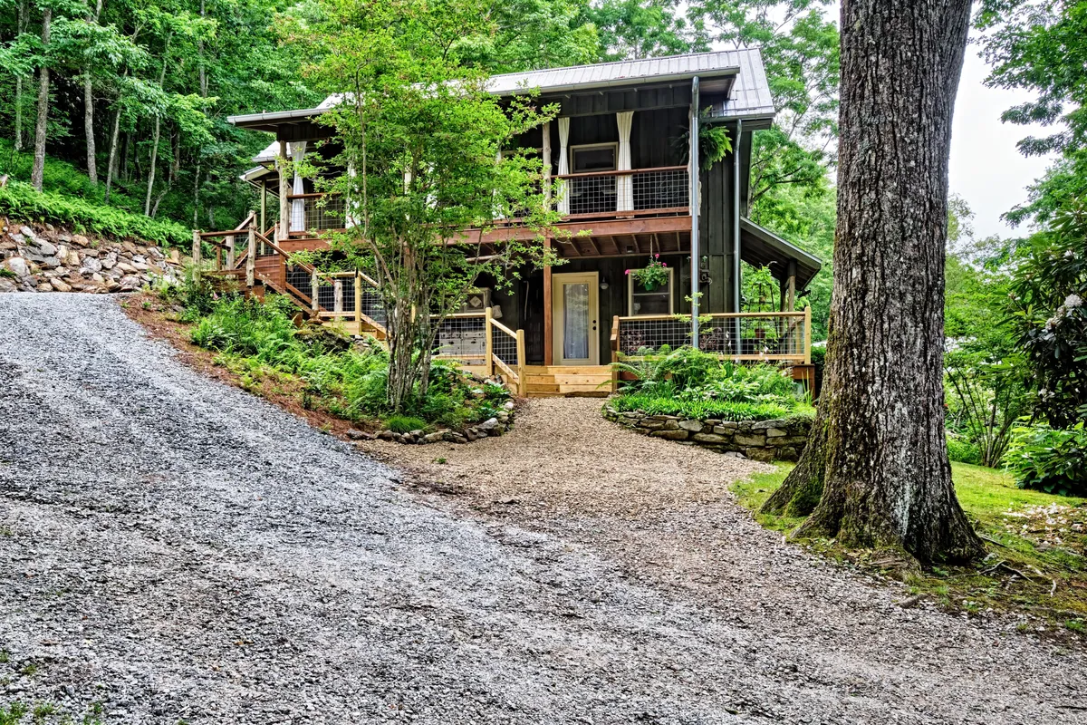 This exterior image showcases the front porch and stone pathway surrounded by lush greenery and a large tree. The setting evokes a sense of privacy and closeness to nature, with the porch offering a welcoming space to relax and enjoy the mountain air. The surrounding woods are apparent, giving context to the property’s secluded atmosphere and proximity to nature trails.