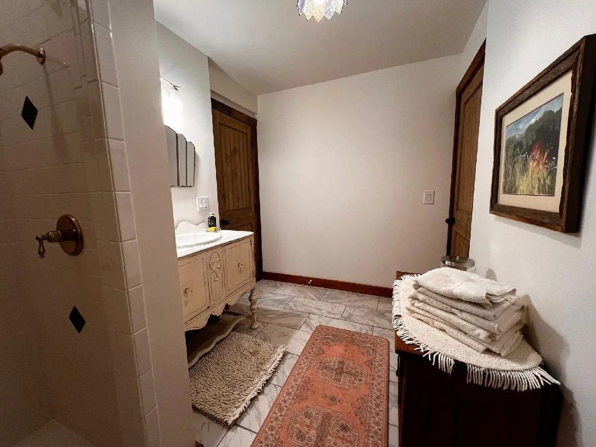 This image features a charming vintage vanity, a shower adorned with decorative tiles, a textured rug, neatly stacked towels, and framed art on the wall. The scene exudes a tranquil and inviting atmosphere, ideal for relaxing bath time or gentle morning routines.