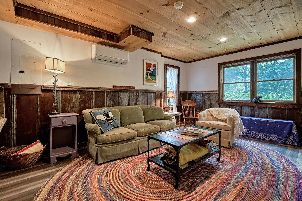 This inviting living room features a comfortable sofa adorned with a whimsical bee pillow, a colorful area rug, and warm wooden walls. Large windows let in natural light, creating a bright, welcoming atmosphere that immediately sets the tone for relaxation and comfort.