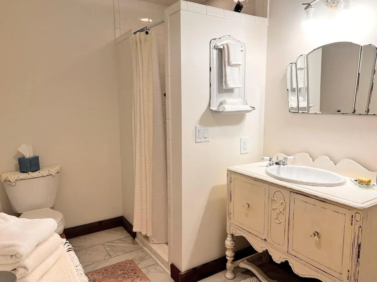 The image showcases a vintage vanity paired with a large mirror, a visible shower area, soft towels, and thoughtfully arranged toiletries. The setting feels serene and cozy, evoking the soothing ritual of a peaceful morning or evening self-care routine.
